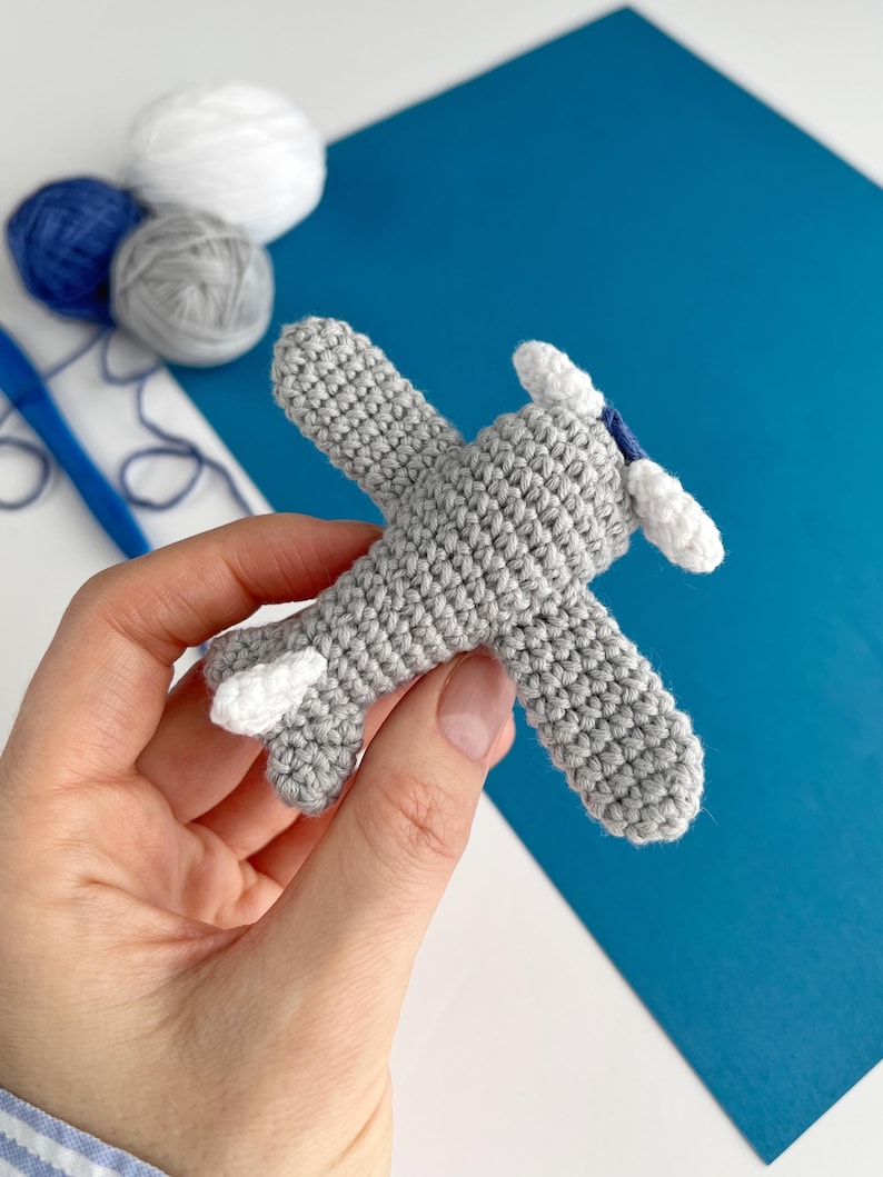 May include: A crocheted gray airplane with white accents on the wings and propeller. The handmade toy is held in a hand, with blue, white, and gray yarn balls in the background. The airplane is a charming, handcrafted item.