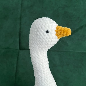 May include: A handmade white crochet goose with a yellow beak and a black eye. The goose is made of soft yarn and is likely a decorative item or a toy. The background is a dark green fabric.