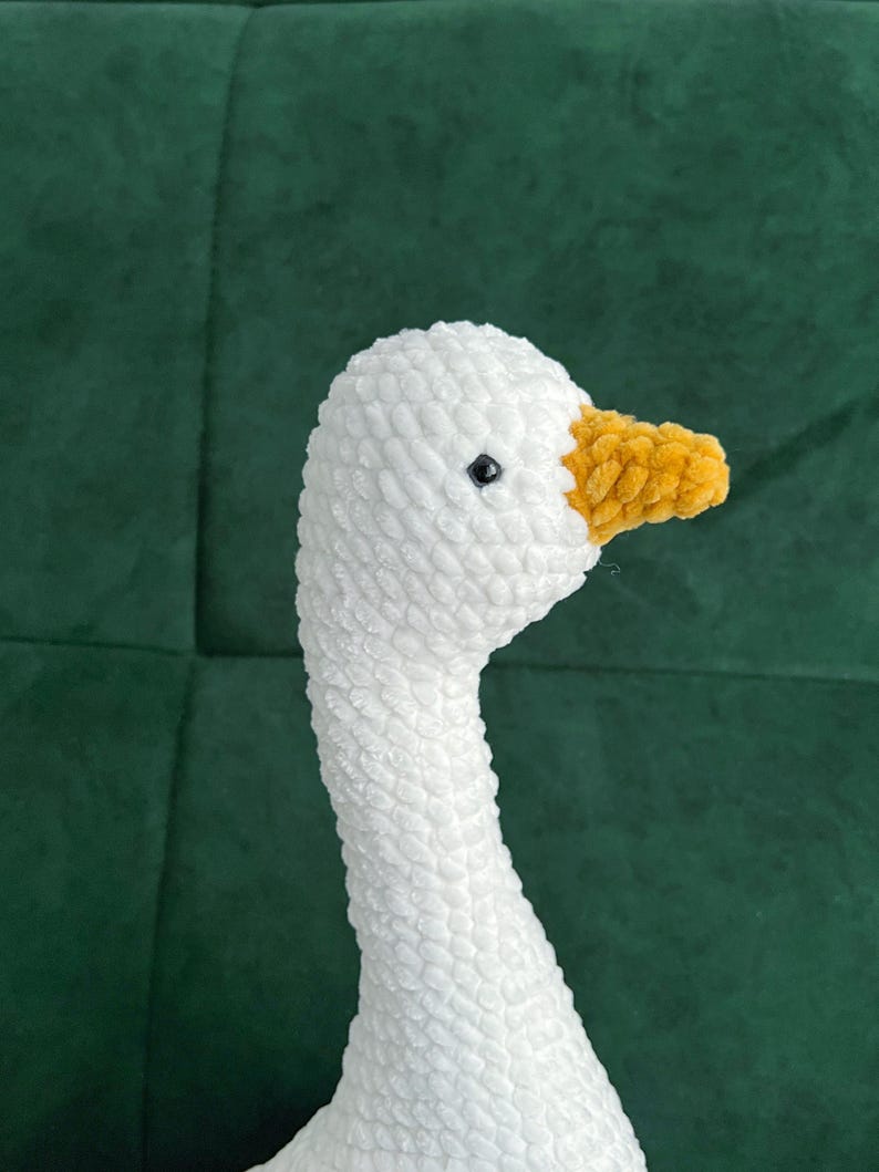 May include: A handmade white crochet goose with a yellow beak and a black eye. The goose is made of soft yarn and is likely a decorative item or a toy. The background is a dark green fabric.