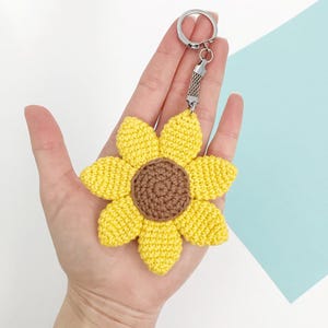 May include: A yellow and brown crochet sunflower keychain with a silver-colored metal keyring and chain. The sunflower has yellow petals and a brown center. The keychain is held in a hand.
