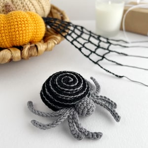 May include: A crocheted spider featuring a round, black and grey spiral-patterned abdomen and a small, solid grey head. The spider has eight articulated grey legs extending outward from the central body, crafted with detailed yarn stitching.