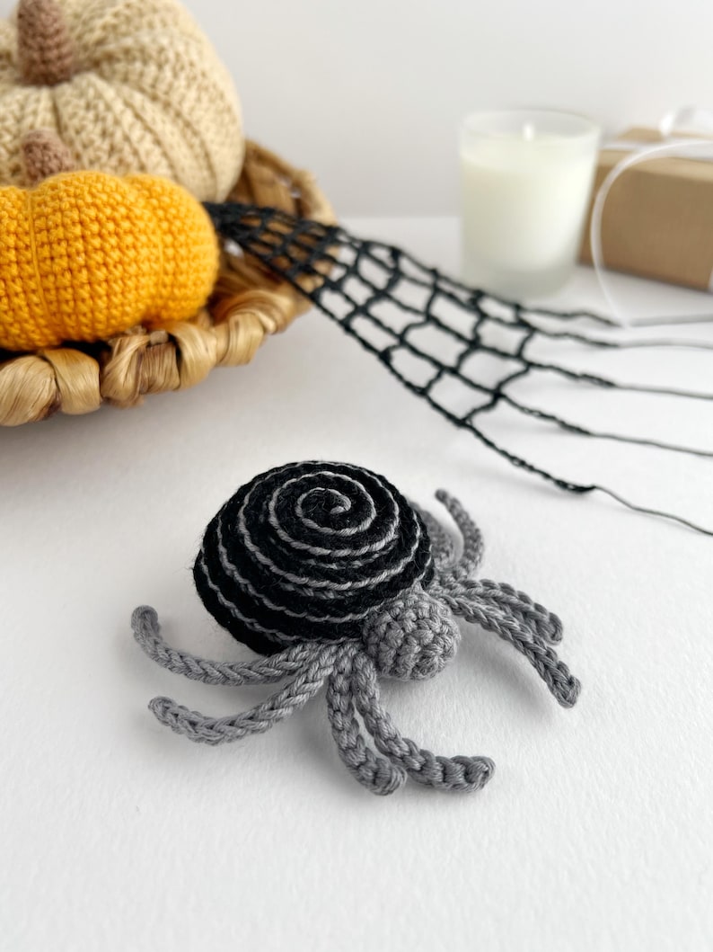 May include: A crocheted spider featuring a round, black and grey spiral-patterned abdomen and a small, solid grey head. The spider has eight articulated grey legs extending outward from the central body, crafted with detailed yarn stitching.