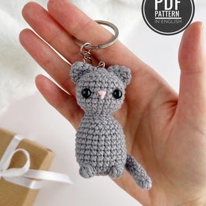 May include: A gray crocheted cat keychain featuring black plastic eyes, a small pink stitched nose, and a metal split ring. The image includes a circular black badge with the white text PDF PATTERN IN ENGLISH.