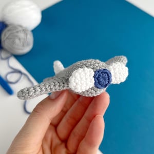 May include: A handmade crochet manta ray toy in gray, white, and navy blue. The toy features a detailed design with rounded wings and a textured body. The manta ray is held in a hand against a blue and white background.