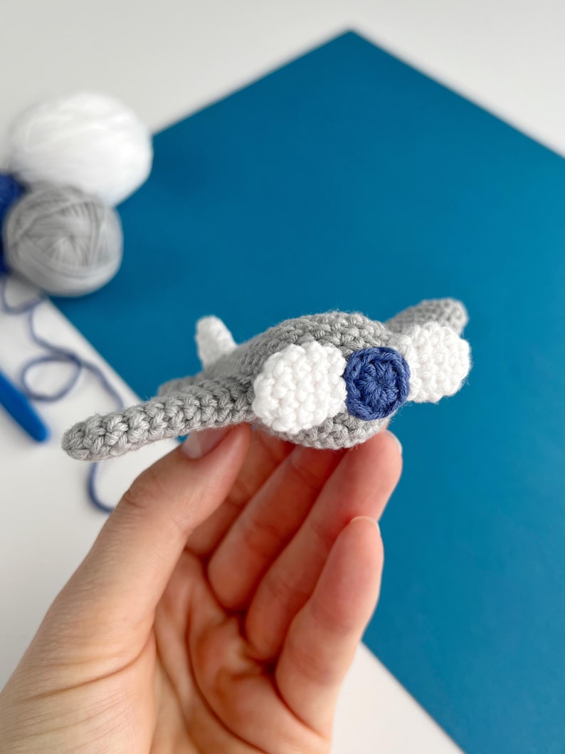 May include: A handmade crochet manta ray toy in gray, white, and navy blue. The toy features a detailed design with rounded wings and a textured body. The manta ray is held in a hand against a blue and white background.