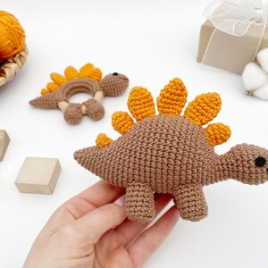 May include: Crocheted dinosaur baby toys in brown and orange. One is a rattle, the other a stuffed animal. The toys are handmade and sit on a white surface. A hand is holding the stuffed animal. A basket of yarn and wooden blocks are in the background.