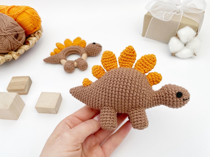 May include: Crocheted dinosaur baby toys in brown and orange. One is a rattle, the other a stuffed animal. The toys are handmade and sit on a white surface. A hand is holding the stuffed animal. A basket of yarn and wooden blocks are in the background.
