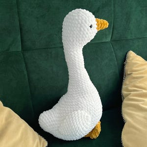 May include: A white, plush goose toy with a long neck and yellow beak and feet. The goose is made of a soft, textured material and is sitting on a green surface. The toy is surrounded by yellow pillows.