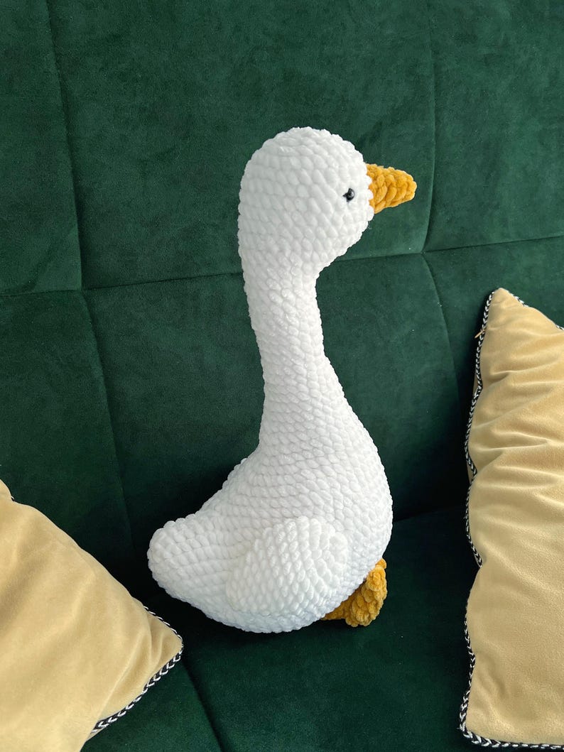 May include: A white, plush goose toy with a long neck and yellow beak and feet. The goose is made of a soft, textured material and is sitting on a green surface. The toy is surrounded by yellow pillows.