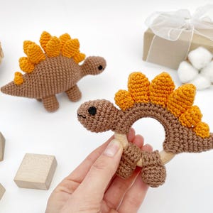 May include: Crocheted dinosaur baby toys in brown and orange. One is a rattle with a wooden ring, and the other is a small dinosaur figure. The toys are handmade and sit on a white surface with wooden blocks and a gift box.