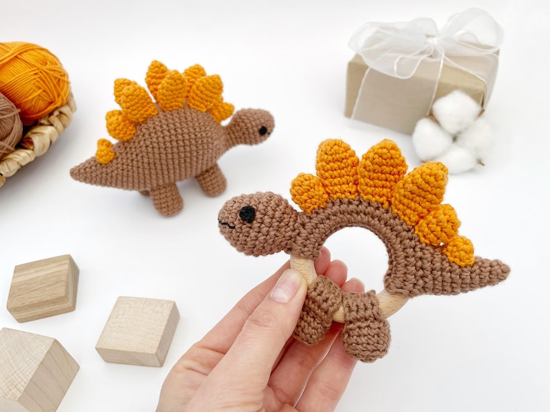 May include: Crocheted dinosaur baby toys in brown and orange. One is a rattle with a wooden ring, and the other is a small dinosaur figure. The toys are handmade and sit on a white surface with wooden blocks and a gift box.