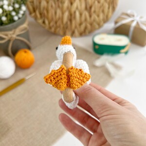 May include: A crocheted goose toy with orange feet attached to a wooden ring. The toy is held in a hand.