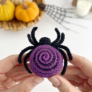 May include: A crocheted spider with a round body featuring a purple and black spiral pattern. The spider has a small black head and eight black crocheted legs extending from the sides. The texture is consistent with yarn craft construction.