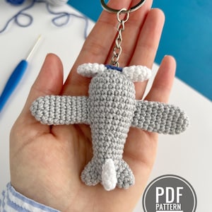 May include: A handmade gray crochet airplane keychain with white accents on the wings and tail. The keychain is attached to a silver metal ring. The image also shows blue and gray yarn and a crochet hook. The text "PDF PATTERN IN ENGLISH" is in a circle.