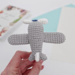 May include: A gray crocheted airplane toy with white and blue details. The toy is held in a hand.