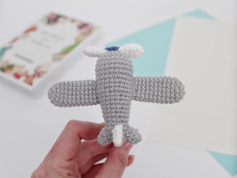 May include: A gray crocheted airplane toy with white and blue details. The toy is held in a hand.
