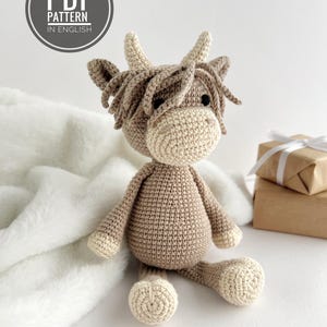 May include: A handmade crocheted Highland cow toy in beige and brown tones. The cow features a light beige face, horns, and hooves, with a darker brown body and fringe. The image includes a circular graphic that reads "PDF PATTERN IN ENGLISH".