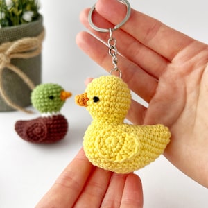 May include: A yellow crocheted duck keychain with a silver keyring. The duck has an orange beak and black eyes. Two other crocheted ducklings, one green and one brown, are visible in the background. The keychain is held in a person's hand.