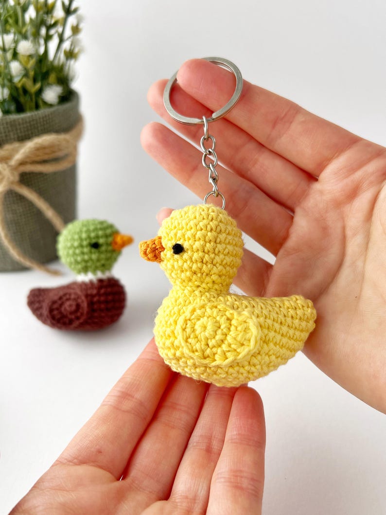 May include: A yellow crocheted duck keychain with a silver keyring. The duck has an orange beak and black eyes. Two other crocheted ducklings, one green and one brown, are visible in the background. The keychain is held in a person's hand.