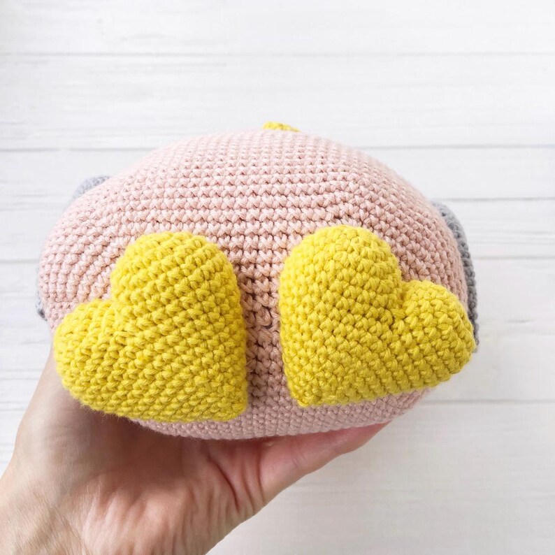 May include: A crocheted stuffed toy in the shape of a bird with pink and gray body and yellow heart-shaped wings.