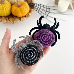 May include: Two crocheted spider ornaments, one featuring a purple spiral body with black legs and the other a black spiral body with gray legs. Each spider has a small round head and eight curved legs extending from the central circular body.