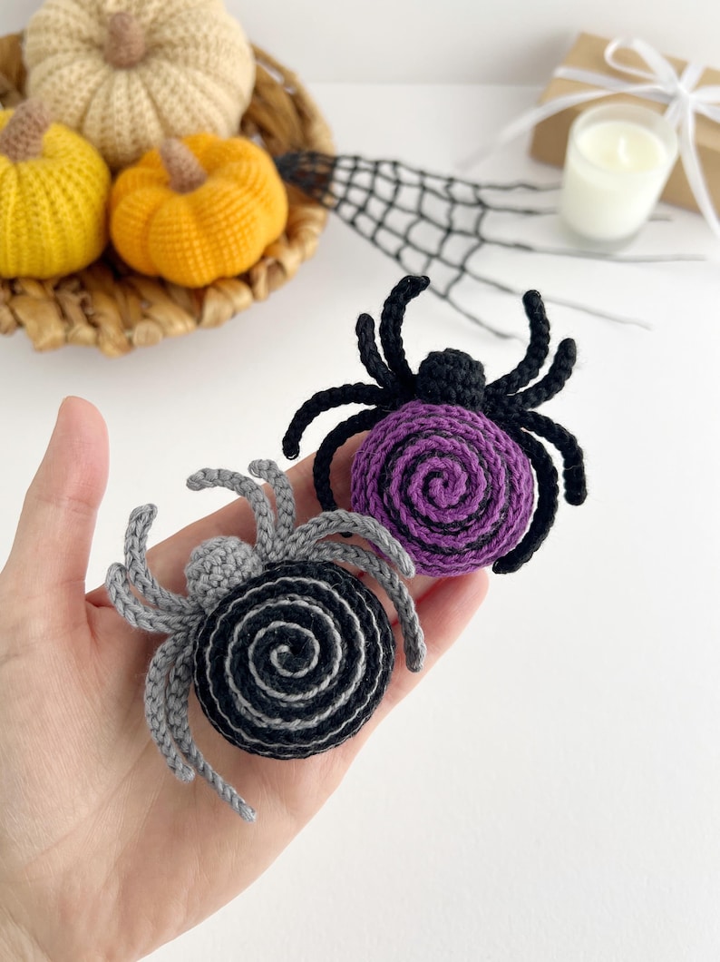 May include: Two crocheted spider ornaments, one featuring a purple spiral body with black legs and the other a black spiral body with gray legs. Each spider has a small round head and eight curved legs extending from the central circular body.