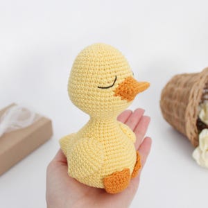 Puede incluir: Un pato amarillo de crochet con pico y patas naranjas. El pato tiene ojos negros y est&aacute; sentado en una mano.