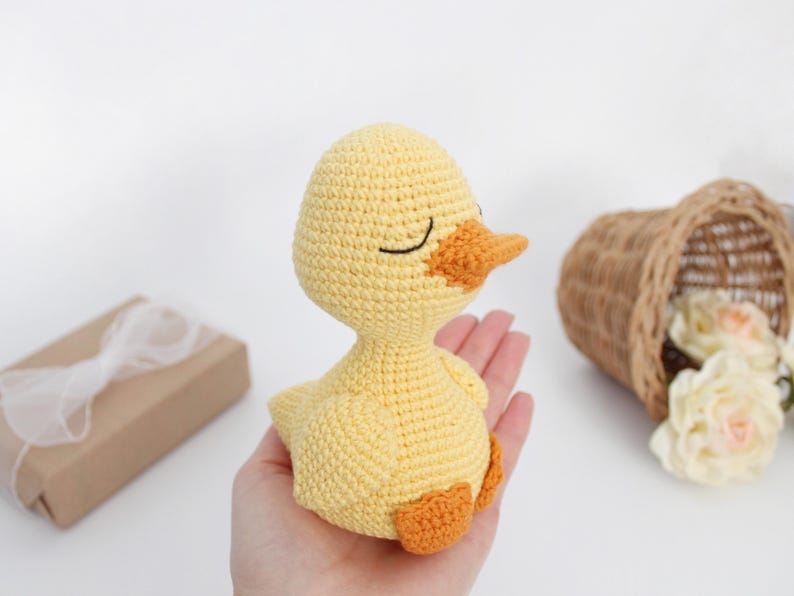 Puede incluir: Un pato amarillo de crochet con pico y patas naranjas. El pato tiene ojos negros y est&aacute; sentado en una mano.