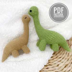 Op de afbeelding: Twee gehaakte dinosaurus speelgoed, één bruin en één groen, liggen op een witte, getextureerde ondergrond. De groene dinosaurus heeft zwarte ogen. Een cirkelvormige afbeelding leest "PDF PATTERN IN ENGLISH".