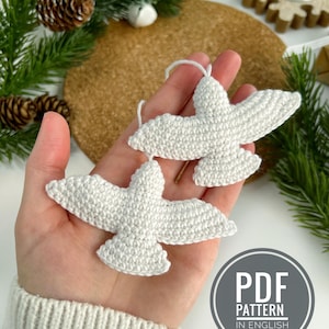 Puede incluir: Dos adornos de paloma de ganchillo blanco con lazos de hilo, sostenidos en una mano. Los adornos tienen forma de pájaros con las alas extendidas. La imagen incluye piñas, vegetación y un gráfico circular que dice "PDF PATTERN IN ENGLISH."