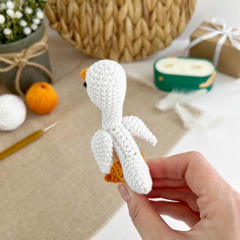 May include: A white crocheted goose with orange feet. The goose is being held in a hand.