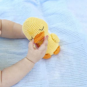 Puede incluir: Un beb&eacute; sostiene un juguete de pato amarillo de crochet con una expresi&oacute;n de ojos cerrados. El pato tiene un pico naranja brillante y est&aacute; acostado sobre una manta de crochet azul.
