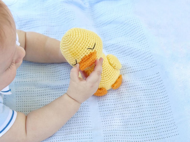 Puede incluir: Un beb&eacute; sostiene un juguete de pato amarillo de crochet con una expresi&oacute;n de ojos cerrados. El pato tiene un pico naranja brillante y est&aacute; acostado sobre una manta de crochet azul.