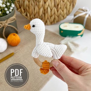 Op de afbeelding: Witte gehaakte ganzenratel met een houten ring en oranje gehaakte voetjes. De ratel wordt in een hand gehouden.