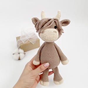 Puede incluir: Un juguete de peluche de vaca de las tierras altas de color marr&oacute;n y crema tejido a crochet con una melena larga y peluda. El juguete est&aacute; siendo sostenido en la mano de una persona.