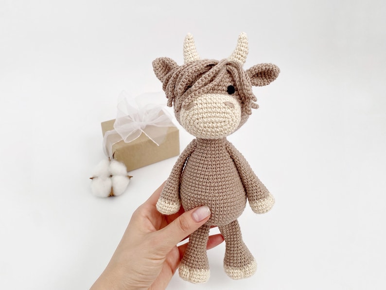 Puede incluir: Un juguete de peluche de vaca de las tierras altas de color marr&oacute;n y crema tejido a crochet con una melena larga y peluda. El juguete est&aacute; siendo sostenido en la mano de una persona.