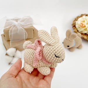 May include: A crocheted beige bunny with a pink bow, sitting in a hand. The bunny has closed eyes and a small nose.