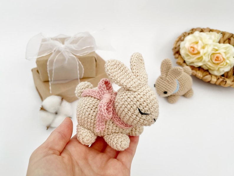 May include: A crocheted beige bunny with a pink bow, sitting in a hand. The bunny has closed eyes and a small nose.