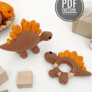 May include: Two crochet dinosaur toys. One is a brown dinosaur with orange spines, and the other is a brown dinosaur-shaped teether with orange spines and wooden accents. The image also includes a ball of orange yarn and a sign that says "PDF PATTERN IN ENGLISH."
