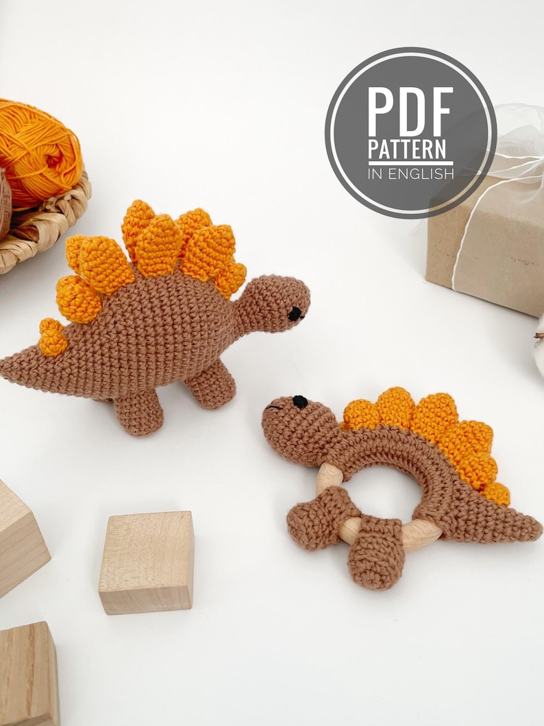 May include: Two crochet dinosaur toys. One is a brown dinosaur with orange spines, and the other is a brown dinosaur-shaped teether with orange spines and wooden accents. The image also includes a ball of orange yarn and a sign that says "PDF PATTERN IN ENGLISH."