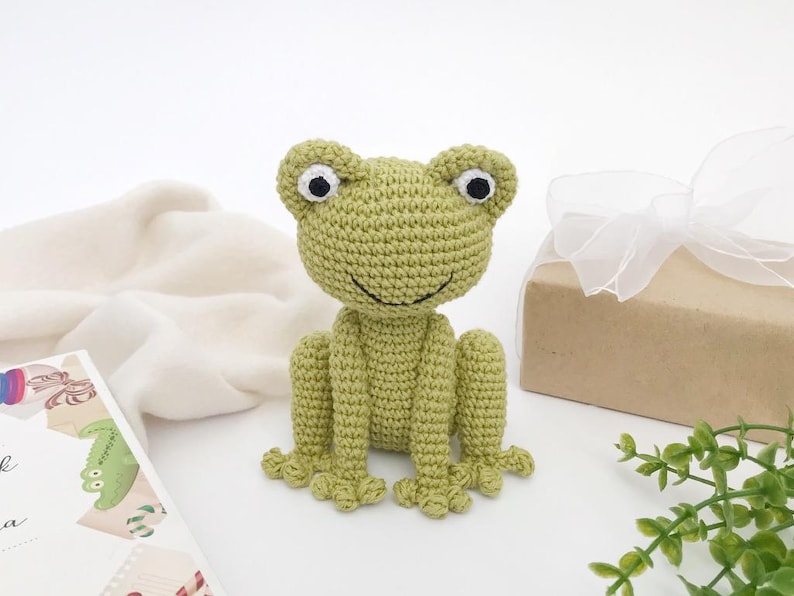 Op de afbeelding: Een handgemaakte, lichtgroene gehaakte kikker met zwart-witte ogen. De kikker zit rechtop met uitgestrekte poten. Een geschenkdoos met een wit lint en een boek staan op de achtergrond.