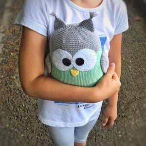 May include: A child holds a crocheted owl plush toy. The owl is green, gray, and white with black eyes and a yellow beak. The child is wearing a white t-shirt with the words "Swim Cool" printed on it.