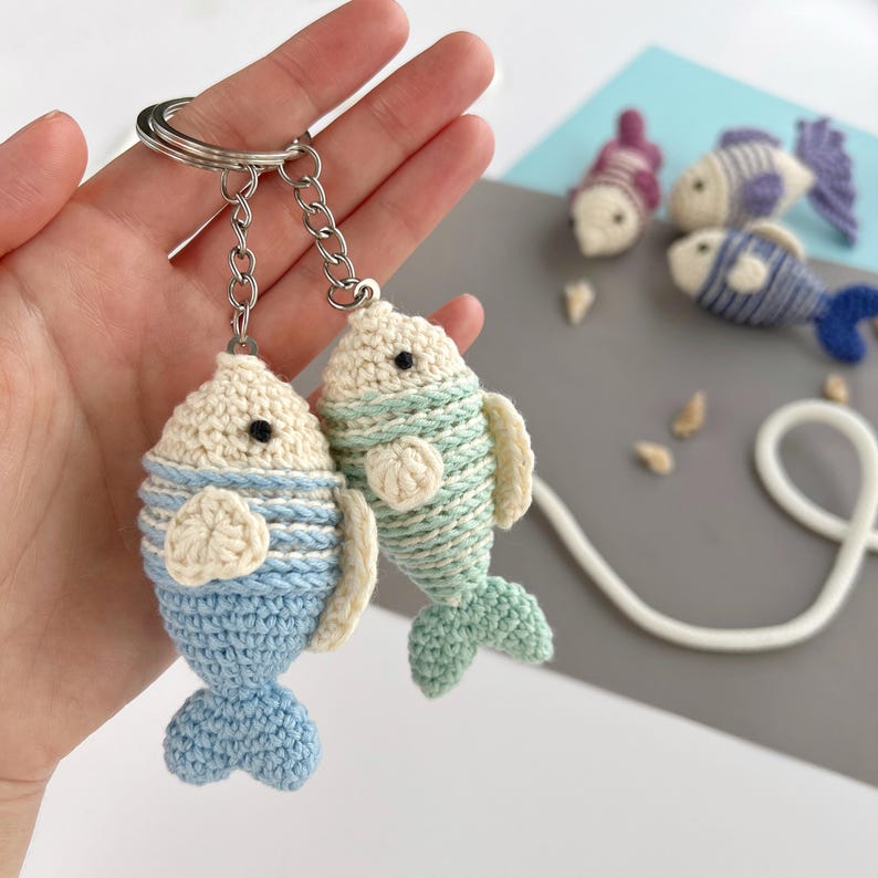 May include: Two handmade crochet fish keychains in blue and green, with cream-colored heads and fins. The keychains are attached to a silver metal ring. Additional crochet fish in the background are in shades of purple and blue.
