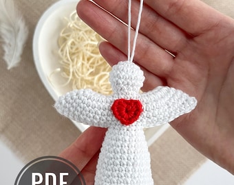 May include: A white crochet angel ornament with outstretched wings and a red heart in the center. The angel is suspended by a white string. The image includes a circular graphic with the text "PDF PATTERN IN ENGLISH".