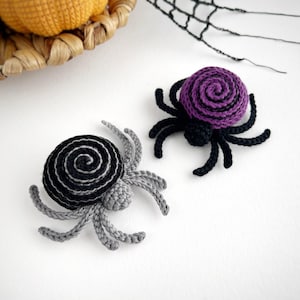 May include: Two handmade crochet spiders, one featuring a black and gray spiral pattern and the other a purple and black spiral pattern. Each spider has eight legs and a small rounded body, crafted with yarn in a detailed, textured stitch.