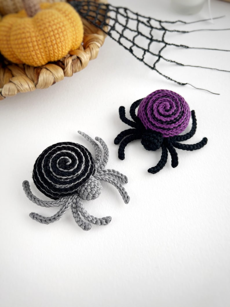 May include: Two handmade crochet spiders, one featuring a black and gray spiral pattern and the other a purple and black spiral pattern. Each spider has eight legs and a small rounded body, crafted with yarn in a detailed, textured stitch.