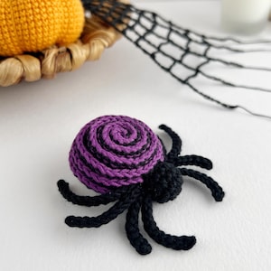 May include: A crocheted spider featuring a round body with a purple and black spiral pattern and eight black legs. The spider is crafted from yarn and has a small, solid black head section attached to the front of the larger, spiraled abdomen.