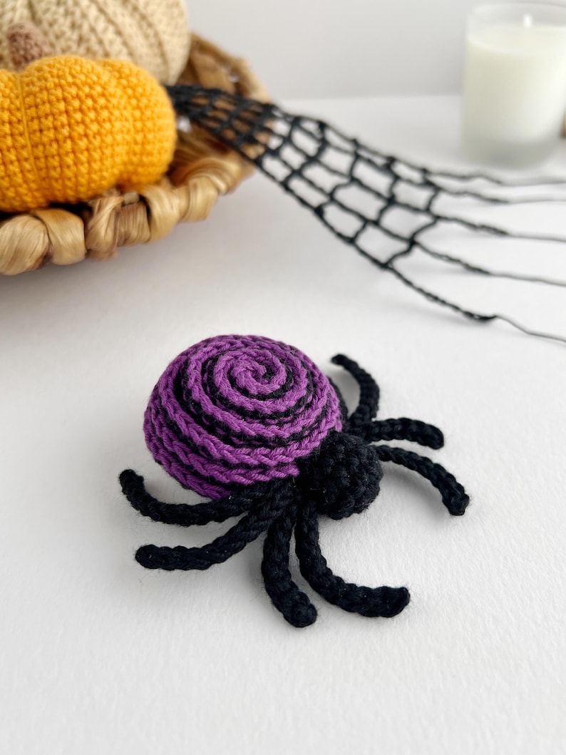 May include: A crocheted spider featuring a round body with a purple and black spiral pattern and eight black legs. The spider is crafted from yarn and has a small, solid black head section attached to the front of the larger, spiraled abdomen.