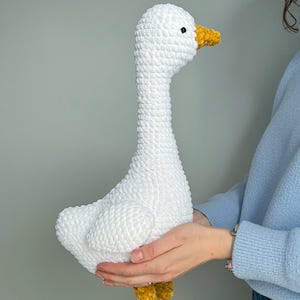 May include: A handmade, white crochet goose with a long neck, yellow beak, and yellow feet. The plush toy is held in front of a light gray background. The goose is a soft, textured, and cuddly decorative item.