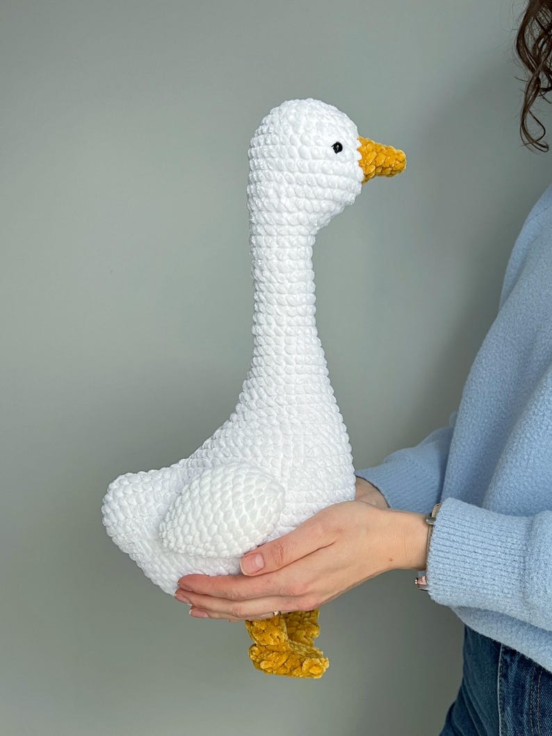 May include: A handmade, white crochet goose with a long neck, yellow beak, and yellow feet. The plush toy is held in front of a light gray background. The goose is a soft, textured, and cuddly decorative item.